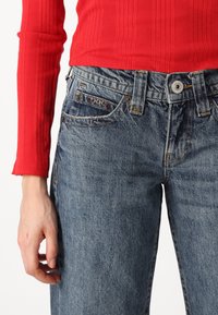 Modré džíny z denimu s stredne vysokým pásom, so zapínaním na gombík a zips, a prednými vreckami. V kombinácii s červeným rebrovaným tričkom s dlhým rukávom.