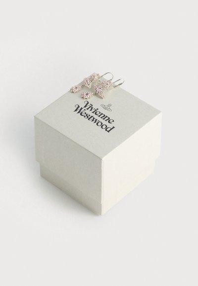 Vivienne Westwood bézs színű négyzet alakú ékszeres doboz fekete logóval; rózsaszín díszített fülbevalók csillag alakúak, lelógó díszekkel, a doboz tetején elhelyezve.
