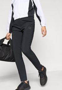 Juodos sportinės kelnės, pagamintos iš lengvos medžiagos, turi lygią apdailą ir subtilų logotipą. Asmuo laiko juodą kelioninę kuprinė eidamas.