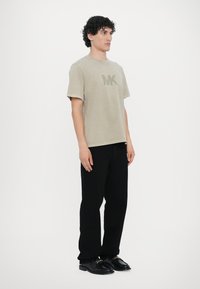 Lys beige bomull t-skjorte med korte ermer og en grønn "MK" logo, paret med svarte bukser og blanke svarte sko.