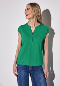 Zelený krátký rukáv top s henley výstřihem, obsahující tři knoflíky a volný střih, ve spojení s modrými džíny.