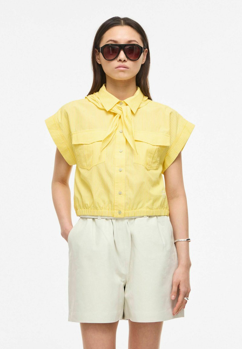Femme portant de grandes lunettes de soleil foncées, une chemise jaune à manches courtes boutonnée avec un col noué, et un short clair taille haute.