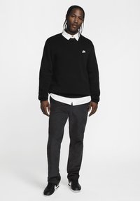 Svart sweatshirt med vitt Nike-logotyp, lager på lager över en vit krag skjorta. Mörkgrå byxor och svarta sneakers, enkel design och texturer.