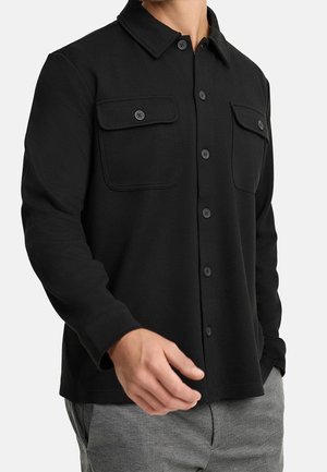 Homme portant une chemise noire boutonnée avec deux poches poitrine et un pantalon gris, visible du cou aux hanches sur un fond uni.