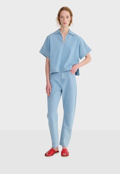 Mujer de pie con una blusa de manga corta y cuello en color azul claro, vaqueros de talle alto a juego y sandalias planas rojas abiertas en un fondo liso.