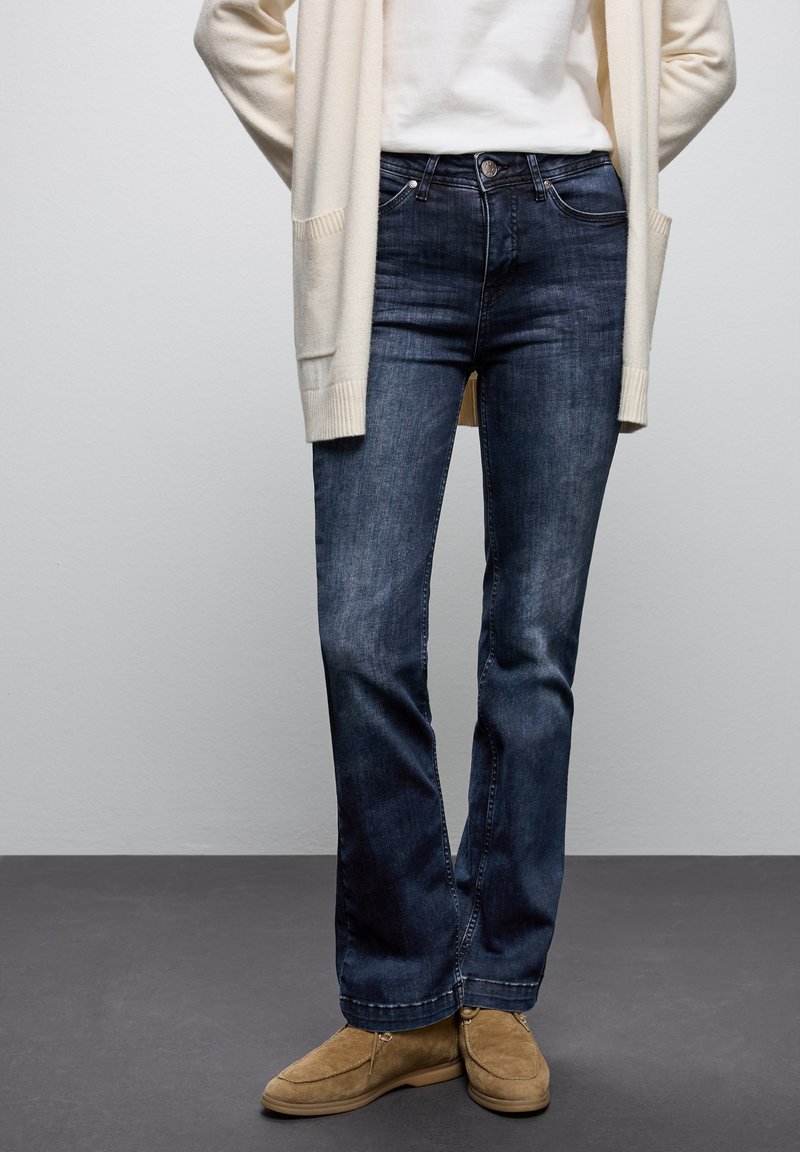 Jeans de denim azul con un corte recto, que presentan un diseño clásico de cinco bolsillos y un ligero desgaste. Combinados con un cárdigan crema y zapatos beige.