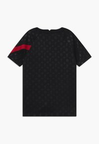 Musta lyhythihainen paita, jossa on hienovarainen tummanharmaa kuvio logoista ja symboleista, ja jossa on vasemmassa hihassa leveä punainen raita.