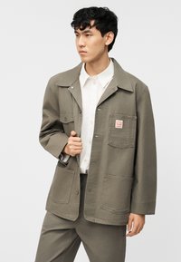 Νεαρός άνδρας που κοιτάζει αριστερά, φοράει χακί μπουφάν με τρεις μεγάλες τσέπες και από μέσα λευκό πουκάμισο με κουμπιά.