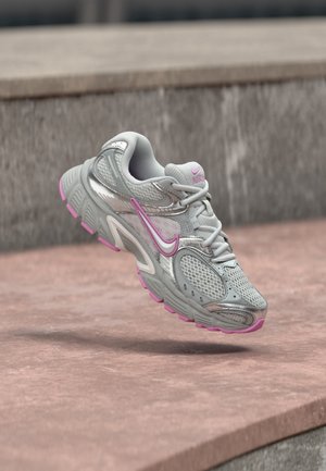 Szaro-różowy but do biegania Nike unoszący się nad różową kamienną powierzchnią z schodami w tle.