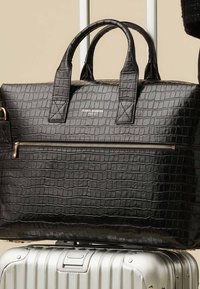 Sac en cuir noir embossé crocodile avec deux poignées, poche zippée à l'avant et garnitures métalliques, posé sur une valise argentée texturée.
