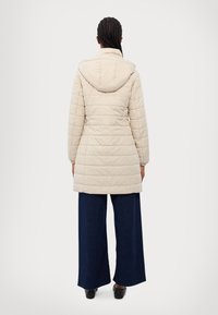 Manteau matelassé beige clair avec capuche, présentant des coutures verticales, des manches longues et une coupe mi-longue. Assorti à un pantalon bleu foncé à jambes larges.
