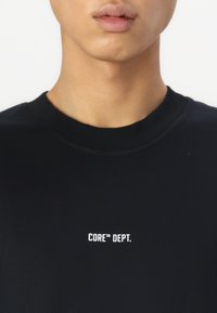 Svart hettegenser med ribbestrikket rund hals. Har hvit tekst "CORE™ DEPT." trykt på brystet. Glatt tekstur, ingen synlige mønstre.