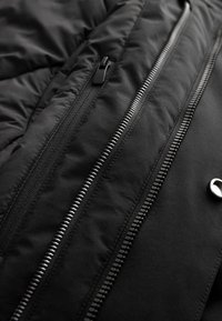 Gros plan sur une veste noire montrant des fermetures éclair parallèles en métal et en plastique ainsi qu'un bouton-pression argenté sur un tissu matelassé.