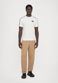 White short-sleeve t-shirt with black accents and logo, paired with tan cargo pants. Model stands on a plain background, wearing black shoes.