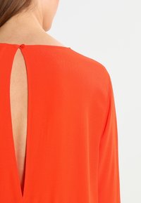 Punainen mekko, jossa on V-muotoinen selkä ja nappikiinnitys. Pehmeä, sileä kangas, jossa on hienovarainen kiilto ja pitkät hihat.