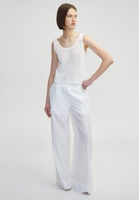Weißes, gestricktes Tanktop mit breiten Trägern, kombiniert mit fließenden, hochgeschnittenen weißen Hosen, die einen leichten Glanz und vordere Falten aufweisen.