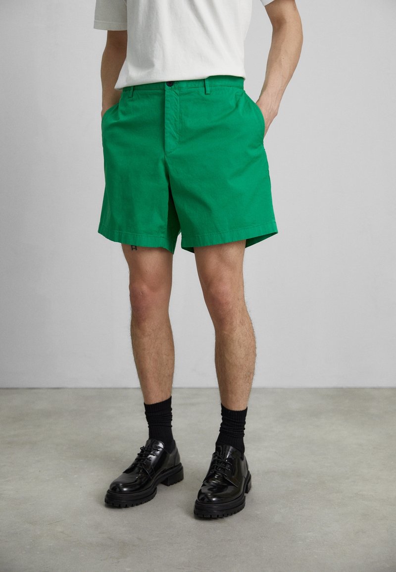 Homme portant un short vert vif, une chemise blanche, des chaussettes noires et des chaussures habillées noires, debout contre un mur et un sol gris.