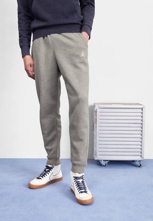Personne portant un pantalon de survêtement Jordan gris, un sweat à capuche bleu marine et des baskets montantes blanches Nike à semelles en caoutchouc, debout sur un sol bleu près d'une boîte de rangement en métal.