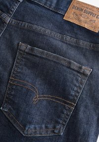 Tumedat sinised denim püksid näevad ette ühe tagatasku, millel on kontrastne heledat pruunist niidist õmblus ja nahkne brändi silt vöökoht. 