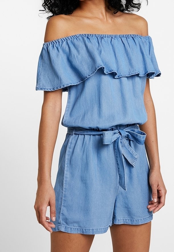 Femme portant une combinaison courte bleue claire à épaules dénudées avec un col à volants et une ceinture en tissu nouée à la taille.