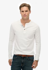 Junger Mann mit kurzen Haaren trägt ein weißes Langarm-Henley-Shirt, blaue Jeans mit braunem Gürtel und eine Armbanduhr, stehend vor einem weißen Hintergrund.