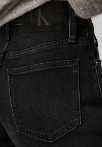 Los vaqueros de mezclilla negra presentan un diseño clásico de cinco bolsillos, costuras sutiles y una superficie texturizada. La cinturilla incluye una etiqueta de cuero negro con el logo.
