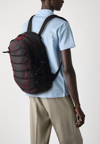 Sac à dos noir avec des accents rouges, plusieurs compartiments, surface texturée et détails en cordon de style bungee. Porté sur l'épaule avec une chemise bleue.