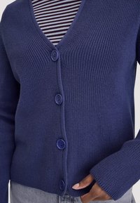 Nahaufnahme einer Person, die eine blaue Strickjacke trägt, die über einem gestreiften Hemd geknöpft ist, während eine Hand den Rand der Strickjacke hält.
