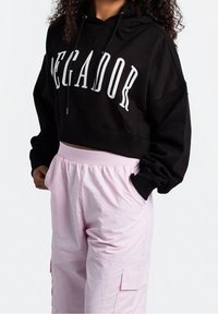 Zwarte cropped hoodie met witte letters "PECADOR," gecombineerd met lichtroze cargobroek met zakken en een elastische tailleband.