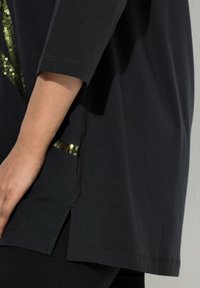 Černé bavlněné tričko s krátkými rukávy, bočními rozparky a detailem ve formě zelených kovových flitrů na přední straně.