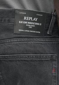 Čierne denimové nohavice s koženým štítkom s nápisom "REPLAY BLUE JEANS MANUFACTURING CO," založené v roku 1981, a červeným prešívaním na zadnej vrecku.