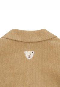 Beiger Cordstoff mit gerippter Textur. Auf der Rückseite des Kragenbereichs befindet sich ein kleines, gesticktes Bärenkopf-Patch.