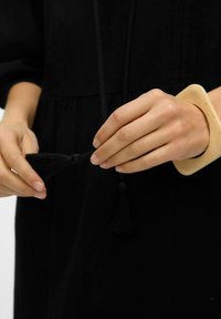 Svart tygplagg med veck, händer som håller en svart tofs-kord och bär ett beige, runt armband i bangle-stil. Texturen ser mjuk ut.