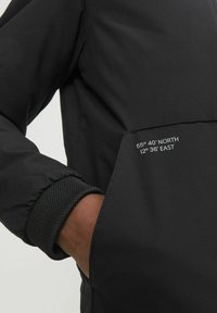 Svart jacka med en dragkedjeficka som har vittryckta koordinater. Ärmsluten har en texturerad stickad detalj. Materialet verkar slätt och elegant.