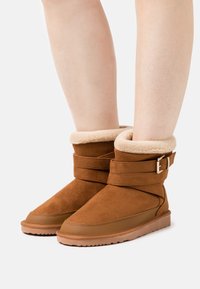 Bruine suède enkelboots met een zachte beige bontvoering, een gewikkelde band met een gouden gesp en een platte rubberen zool.