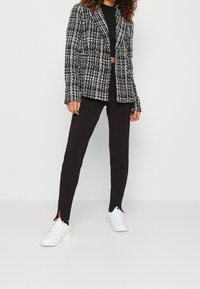 Černobílý tvídový blazer se strukturovanými rameny, předními kapsami a otevřeným designem, doplněný o černé slim kalhoty a bílé tenisky.