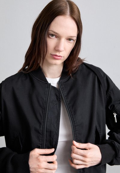 Fekete bomberdzseki cipzárral elöl, bordás mandzsettákkal és oldalsó zsebekkel. Fehér, bordás felső fölött viselve, amely sima és matt textúrájú.