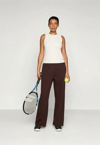 Top sin mangas blanco, pantalones anchos marrones, zapatos negros. La modelo sostiene una raqueta de tenis y una pelota de tenis amarilla, de pie sobre un fondo liso.