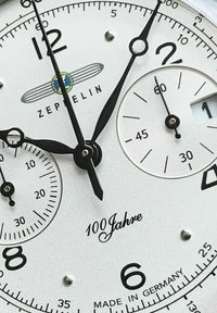 Zeppelin-Armbanduhr mit einem weißen Zifferblatt, schwarzen Stunden- und Minutenzeigern, zwei kleinen Zifferblättern und dem Text "100 Jahre", versehen mit eleganten schwarzen Akzenten.