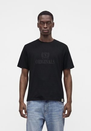 Jeune homme portant un t-shirt noir avec le texte "ORIGINALS" et un jean bleu clair, debout devant un fond blanc uni.