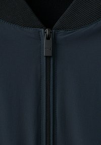 Zbliżenie na suwak granatowej kurtki z czarnym uchwytem oznaczonym "MNG" oraz szczegóły prążkowanego kołnierza.