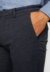 Tmavě šedé kostkované kalhoty s texturovanou tkaninou, vybavené bočními kapsami a kontrastním modrým detailem na pásku.