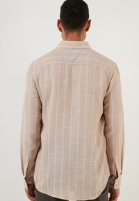 Světle hnědá košile s dlouhými rukávy, tenké bílé vertikální pruhy, límec na knoflíky a spodní část mírně zaoblená po stranách.