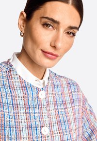 Textur-reicher, mehrfarbiger Tweed-Jacke mit weißem Kragen und zwei großen weißen Knöpfen, aus einer Nahaufnahme der oberen Bekleidung.