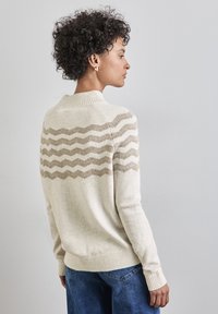 Cremfarbener Pullover mit einem wellenförmigen beigen Muster auf dem Rücken, gerippter Kragen und Bündchen, kombiniert mit blauen Jeans. Strukturiertes Strickgewebe.