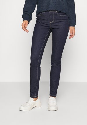 Personne portant un jean skinny bleu foncé, un pull foncé et des chaussures blanches sans lacets, debout devant un fond uni.