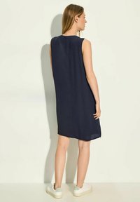 Vestido sin mangas en tejido ligero, de corte relajado, a la altura de la rodilla con aberturas laterales. Combinado con zapatillas blancas con un acento en verde claro.