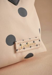 Bežinė audinio rankinė su didelėmis tamsiai pilkomis taškelėmis. Prieduose yra siuvinėta etiketė su tekstu "Cath Kidston" auksine spalva. Lygi tekstūra.