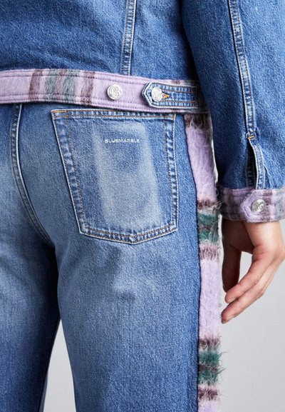 Veste en denim au tissu bleu clair, avec une bordure en peluche rayée lavande et verte le long du col et des poignets. Du matériel en argent est visible.