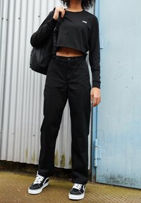 Musta lyhythihainen cropped-paita, jossa on valkoinen logo, yhdistettynä korkeavyötäröisiin mustiin culottes-housuihin ja mustiin lenkkitossuihin, joissa on valkoisia yksityiskohtia. Musta reppu.